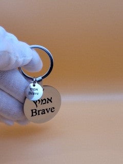 Left hand in white cotton glove holding stainless steel round keychain engraved with word  " Brave" in hebrew and english. 