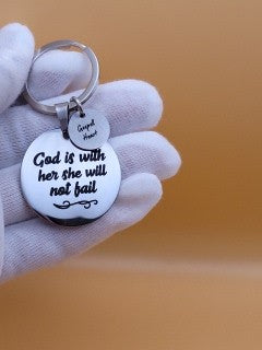 left hand in white cotton glove with keychain on index finger holding stainless steel 35mm round keychain engraved with word  "God is with her she will not fai"